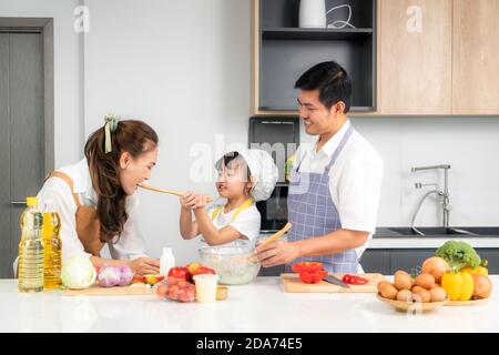 Asian daughters feeding salad to her mother and her father stand by when a family cooking in the kitchen at home. Family life love relationship, or ho Stock Photo