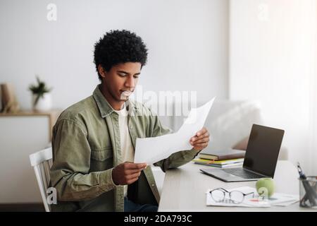 Black teenager studying remotely on laptop, working on coursework, coordinating his school project with tutor at home Stock Photo