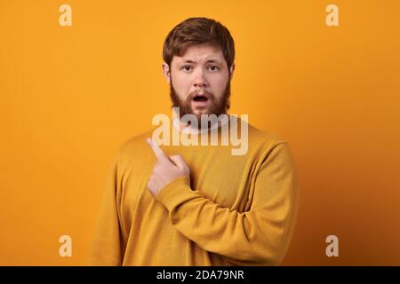 Close up shot of emotive shocked bearded young male opens mouth widely, stare at camera, points upwards aside at blank space, says: Wow, it s Stock Photo
