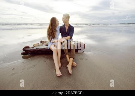 Couple sitting on a dead tree trunk on a beach in Bali at sunset, looking at each other Stock Photo