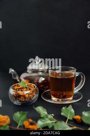 Organic chaga tea on a black background. Infusion with birch mushroom in a glass cup and teapot. Healthy beverage. Vertical orientation. Copy space. Stock Photo