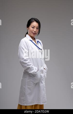 Thoughtful female Asian doctor or nurse in lab coat standing sideways looking t the camera with a quiet friendly smile over a grey studio background w Stock Photo