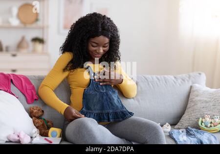 Excited pregnant black woman looking at small dress for baby Stock Photo