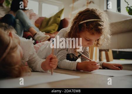 Portrait of two daughter lying on floor making artwork on paper at home with parents sitting on couch Stock Photo