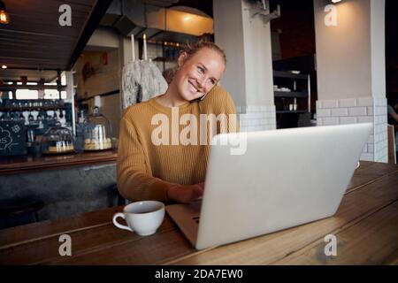 Cheerful young female businesswoman sitting in modern cafe talking over phone and using laptop Stock Photo