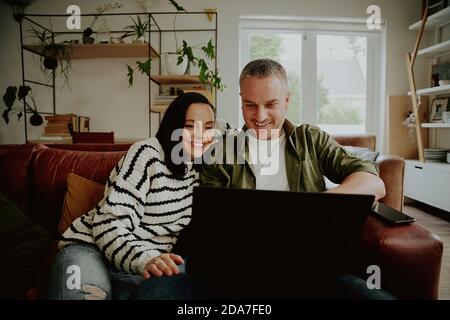 Smiling man and woman using laptop at home while sitting together on sofa Stock Photo