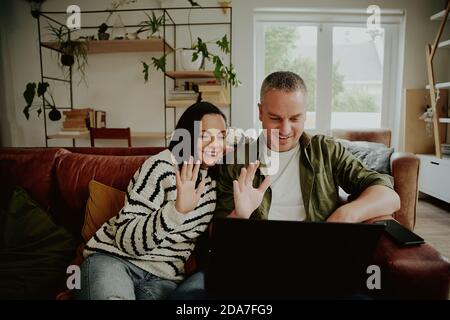 Cheerful couple sitting on sofa and waving hands to laptop while chatting using video call at home Stock Photo