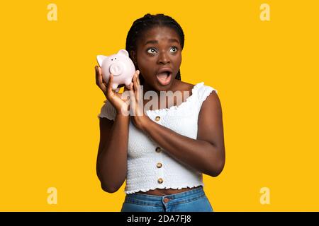 Shocked african american woman listening sound of coins in piggy bank Stock Photo