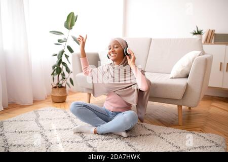 Positive black woman in hijab sitting on floor, wearing headphones, listening to music at home Stock Photo