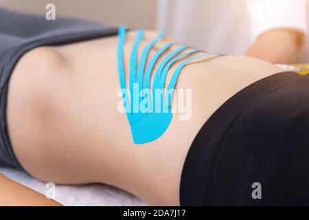 Male physiotherapist applying blue adhesive elastic therapeutic tape on female abdominal muscle of young adult slim sporty woman lying on couch doctor Stock Photo