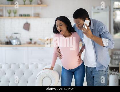 Pregnant African American woman having prenatal contractions, worried husband calling doctor on smartphone Stock Photo