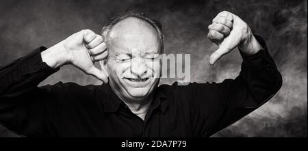 Old macho man gesturing with thumb pointing down. Portrait of an elderly emotional man on a dark background in the studio. Black and white image Stock Photo