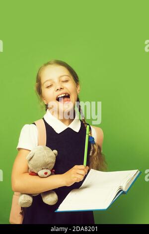 Kid in school uniform isolated on green background. Study and back to school concept. Pupil holds blue book, marker and teddy bear. Girl with braids with open mouth and closed eyes Stock Photo