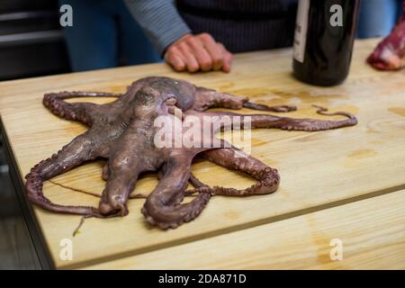 Whole raw octopus lying on wooden board in kitchen ready to be cooked with human hands and wine bottle in background Stock Photo