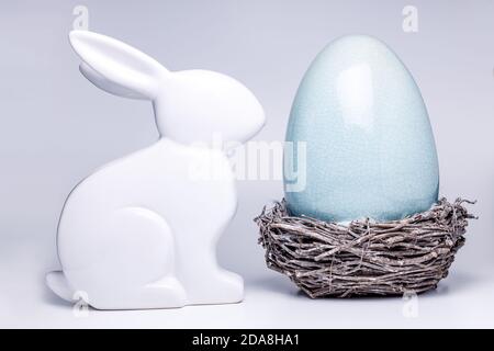 Side view of a white ceramic easter bunny figure besides a easter nest made of twigs and branches with a turquoise mint green ceramic easter egg insid Stock Photo