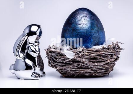 Silver metalic easter bunny figure looks into a royal blue easter egg laying in a easter nest made of brown branches, filled with white feahters on wh Stock Photo