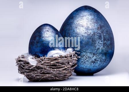 Two royal blue easter eggs in different sizes on a white and uniform background, one egg is in a easter nest with some white and fluffy feathers, popu Stock Photo