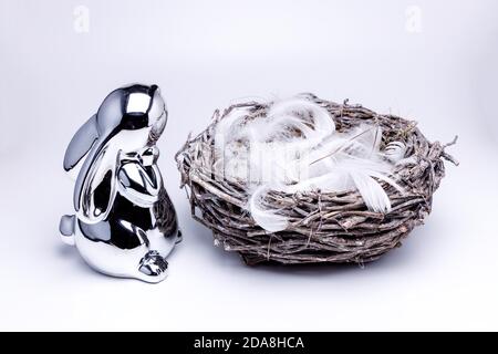 Small and cute silver easter bunny figure looking into a empty easter nest made of branches, filled up with white feathers, white and uniform backgrou Stock Photo