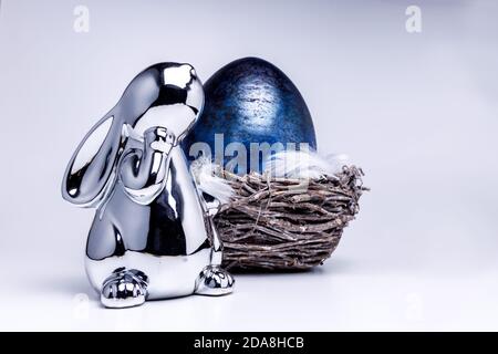 Cute silver metallic easter bunny figure looks onto a royal blue easter egg laying in a easter nest made of brown branches, filled with white feathers Stock Photo