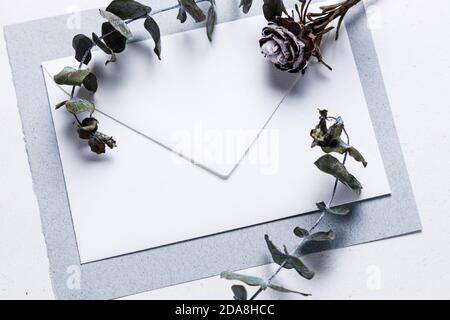 Small white envelope on grey and light grey natural paper, decorated with eucalyptus twigs in soft light, use as wedding card template or background Stock Photo