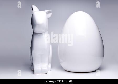 Frontal shot of two easter decoration items, showing a white ceramic figure a easter bunny and a large white egg on a soft white and uniform backgroun Stock Photo