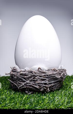 White shiny ceramic easter egg in an easter basket with bird feathers on green artificial lawn for Easter as a background or template, side view Stock Photo