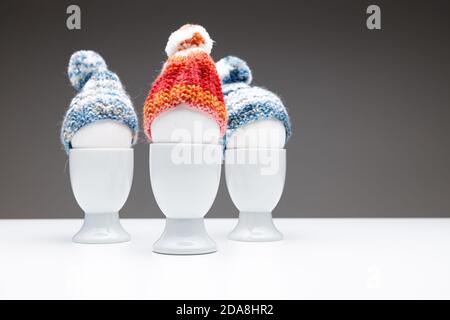Three egg cups with white eggs and wool beanies, on dark grey background on a white table., use for easter greeting card or christmas greeting card. Stock Photo