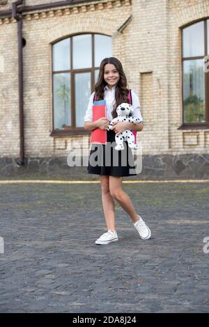 Happy small kid in school uniform hold books and toy dog in schoolyard, homework club. Stock Photo
