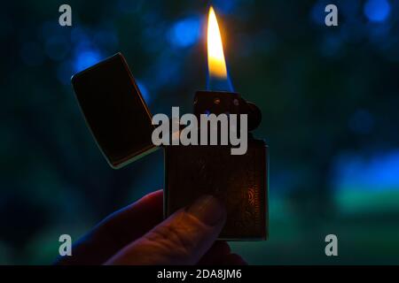 A hand holds a Venetian gold Zippo lighter open and lit. Zippo lighters are reusable metal lighters manufactured by American Zippo Manufacturing. Stock Photo