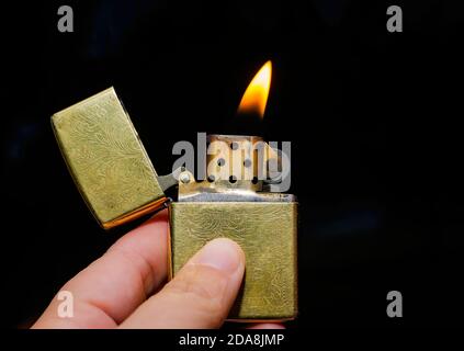 A hand holds a Venetian gold Zippo lighter open and lit. Zippo lighters are reusable metal lighters manufactured by American Zippo Manufacturing. Stock Photo