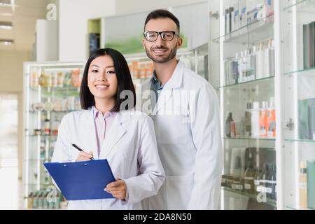 happy interracial pharmacists in white coats looking at camera in drugstore Stock Photo