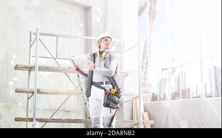 woman worker builder work with digital tablet, wearing helmet, hearing protection headphones and bag tools, on scaffolding construction site indoors b Stock Photo