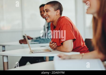 Smiling female student sitting at desk with laptop in high school classroom.  Girl with short hair smiling during the lecture in university classroom. Stock Photo