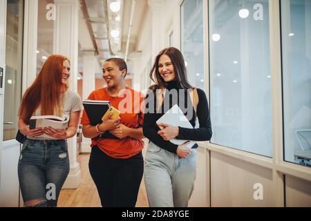 Smiling girls walking through high school corridor after their class. Three female college students after lecture. Stock Photo