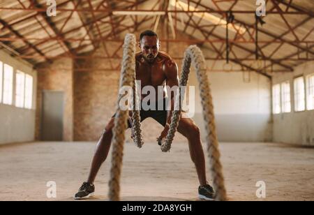 fitness man exercising with battling rope in cross training gym inside old warehouse. Muscular man working out at empty warehouse. Stock Photo