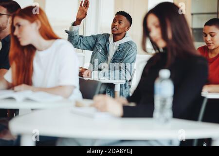 Male student sitting in the class and raising hand to ask a question during lecture. College student asking a query to the lecturer in classroom. Stock Photo