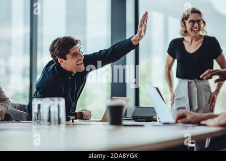 Smiling businessman giving a high five to his coworker sitting around the table in meeting room. Office colleagues sitting in conference room celebrat Stock Photo