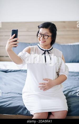 Joyful young pregnant woman sitting on bed and taking selfie on smartphone Stock Photo