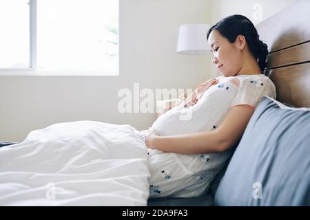 Smiling pretty pregnant young woman sitting in bed and touching belly in the morning Stock Photo
