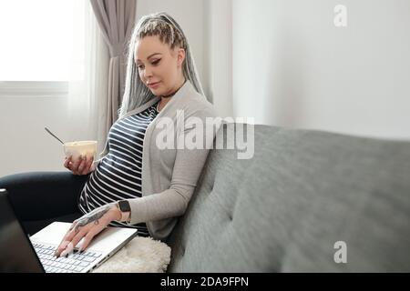 Young pregnant woman having bowl of fruit salad and searching information on maternity online on laptop screen Stock Photo