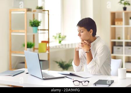 Pensive woman sitting at desk with laptop and notebook watching educational video or reading ebook Stock Photo