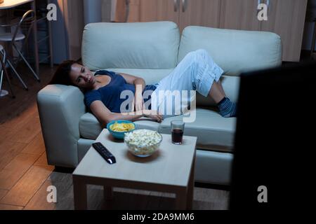 Tired, bored, unhappy housewife using remote control lying on couch laughing and eating snacks. Lonely lady in pijamas enjoying the evening sitting on comfortable sofa watching television. Stock Photo