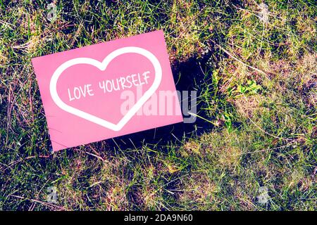 Love yourself concept written on paper with heart shape on green grass background. Inspirational quote for motivation, happiness or success in relatio Stock Photo
