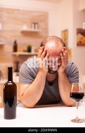 Man having emotional problem sitting alone in kitchen with a glass of red wine on table. Unhappy person disease and anxiety feeling exhausted with having alcoholism problems. Stock Photo