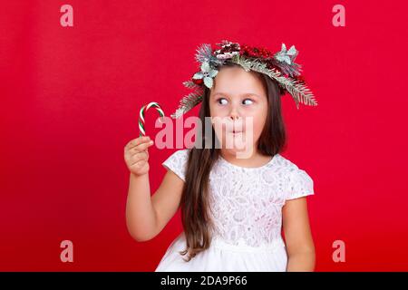 mock up of a beautiful girl in Christmas wreath looking with surprise at caramel cane in her hand, on a red background Stock Photo