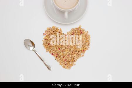 Breakfast. Half a white Cup and saucer of cappuccino. Muesli with red dried berries in the form of a heart and a spoon. Top view, white background. Fr Stock Photo