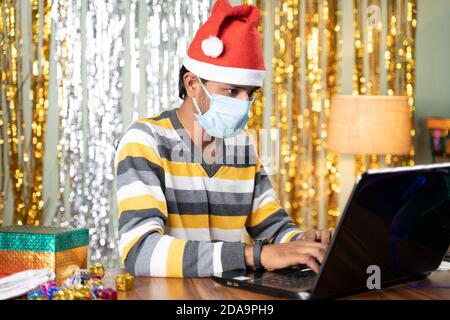 Young man in medical mask busy working on laptop during Christmas or new year celebration eve with decorated background and gifts in front - concept Stock Photo