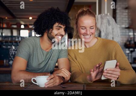 Cheerful young woman showing funny video in smartphone to boyfriend while relaxing and sitting in modern cafe Stock Photo