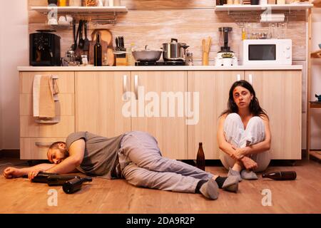 Alcoholic man lying on the floor near beaten wife surrounded by empty bottles. Drunk aggressive husband abusing injuring terrified helpless, afraid, beaten and panicked woman living in terror. Stock Photo