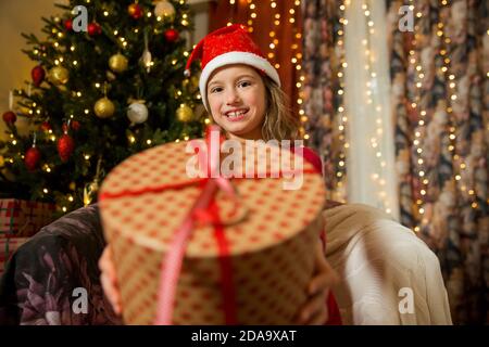 A happy child in red Santa hat is celebrating Christmas. Cute girl holding wrapped gift, smiling and waving hand, sitting in decorated with Christmas Stock Photo